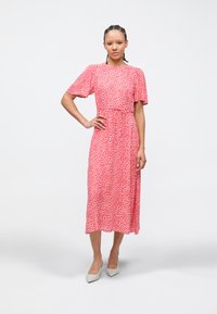 Ροζ μεσαίο φόρεμα με λευκά πουά, κοντά μανίκια σε στυλ φλάτερ, ζωνάκι στη μέση, αέρινη φούστα, συνδυασμένο με ανοιχτό γκρι ψηλοτάκουνα παπούτσια με μυτερή μύτη.