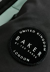 Čierna tkanina s textúrovanou, obručou so zaoblenými okrajmi, na ktorej je nápis "UNITED KINGDOM BAKER BY TED BAKER LONDON" bielym a strieborným písmom.