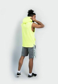Hombre ajustando la capucha de una camiseta sin mangas de color amarillo neón, vistiendo pantalones cortos grises con texto lateral y zapatillas negras, de pie contra un fondo sencillo.