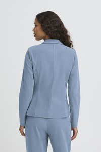 Femeie cu păr lung și creț, privită din profil, purtând un sacou albastru deschis mulat și pantaloni asortați, pe un fundal simplu.