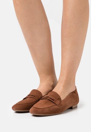 Mocassins en suede marron avec un bout arrondi, présentant une sangle décorative à l'avant et un petit talon carré. Texture lisse, design décontracté.