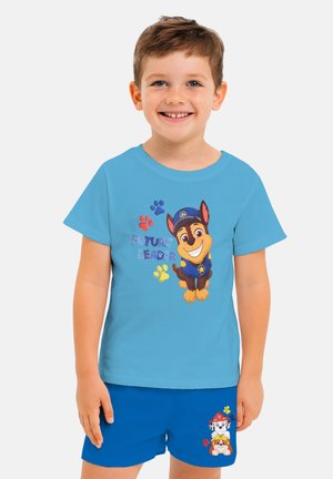 Helles blaues T-Shirt mit einem Cartoon-Hund in einer Kappe, dem Text "ZUKÜNFTIGER FÜHRER" und bunten Pfotenabdrücken. Kombiniert mit blauen Shorts, die Hundepersönlichkeiten zeigen.