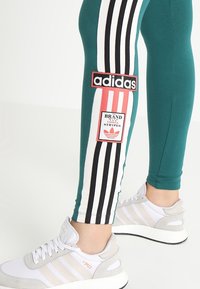 Benen in turquoise leggings met zwart-witte strepen en Adidas-logo's, gecombineerd met witte Adidas-sneakers met beige accenten.