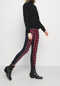 Personne marchant en col roulé noir, bottes à la cheville noires et pantalon slim-fit avec des panneaux avant à carreaux rouges et des panneaux arrière bleu foncé.