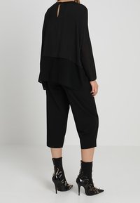 Blusa nera a strati con retro a chiave, abbinata a pantaloni neri a tre quarti e tacchi aperti con accenti a punta.