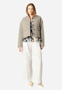 Gepatchte graue cropped Jacke mit Knopfverschluss, getragen über einer blumigen Bluse, kombiniert mit weißen weit geschnittenen Hosen und beigen Schuhen.