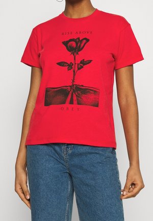 Persoon draagt een rood T-shirt met een zwarte rozenafbeelding en de tekst "RISE ABOVE" boven en "OBEY" onder, gecombineerd met een blauwe spijkerbroek.