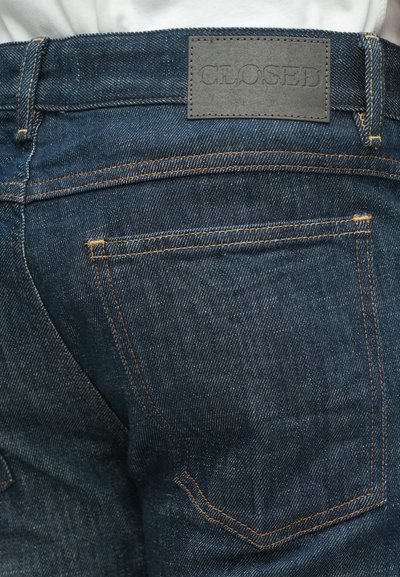 Jean en denim bleu foncé avec un patch en cuir marqué "CLOSED." Caractérisé par des coutures orange et deux poches arrière avec une usure minimale.