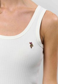 Bela rebrasta majica brez rokavov z majhnim vezenim motivom na levi strani prsnega koša, ki prikazuje teksturirano blago in prilegajočo silhueto.