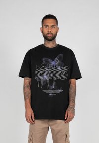 Svart oversized t-skjorte med grafisk design av gjennomsiktige blå sommerfugler og stilisert tekst. Korte ermer og rund hals.
