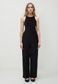 Jonge vrouw staat tegen een effen achtergrond in een mouwloos zwart topje, gestructureerde zwarte wijde broek en zwarte platte schoenen.