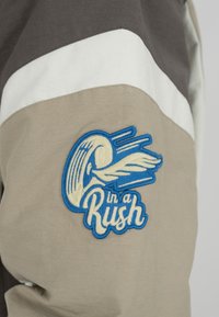 Tissu beige, blanc et marron avec un patch brodé bleu et crème représentant une vague ailée et les mots « in a Rush ».