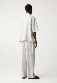 Homme à la peau foncée portant une chemise ample en lin beige clair et un pantalon large, debout de profil avec des sandales noires à boucle sur fond blanc.