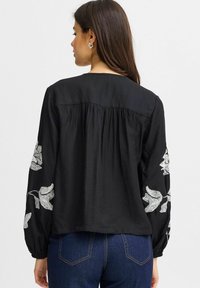 Blusa nera con maniche lunghe e ampia, caratterizzata da ricami bianchi a forma di foglia. Tessuto morbido e vestibilità ampia con dettaglio arricciato sul retro.