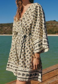 Robe de couleur claire avec un imprimé floral et des accents noirs. Présente un décolleté en V, des manches larges et un lien à la taille. Tissu texturé.