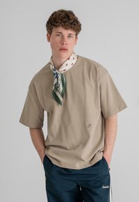 Beige, rövid ujjas póló, kerek nyakkivágással, amelyen egy kicsi logó található a mellkason. Egy színes mintás sál kíséri, amely a nyak köré van kötve.