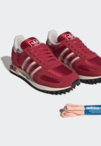Paire de baskets rouges Adidas avec trois bandes rose clair, tissu texturé et lacets roses supplémentaires à côté sur fond blanc.