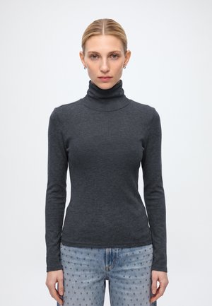 Femme aux cheveux clairs portant un pull à col roulé gris foncé et un jean bleu clair avec de petits clous, debout devant un fond blanc uni.