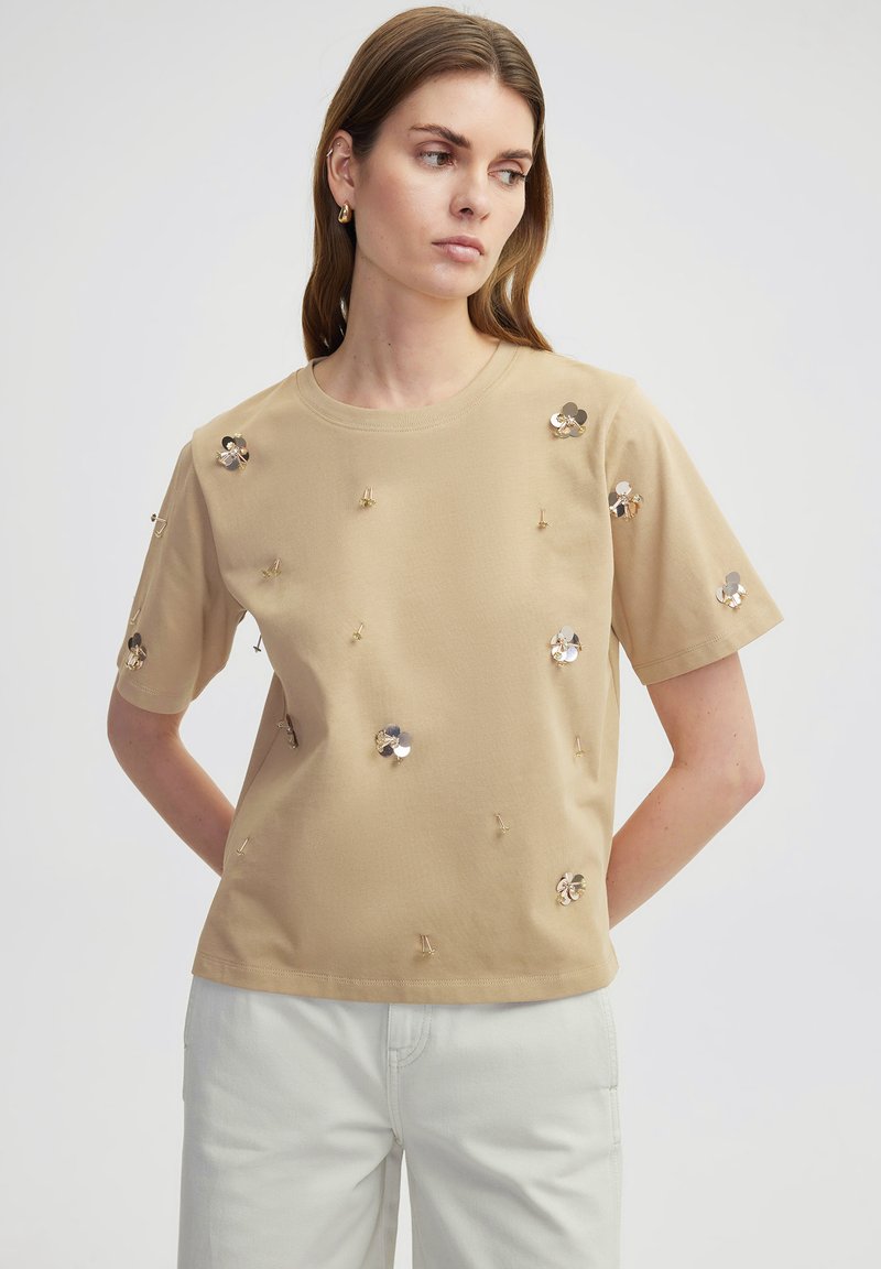 T-shirt in cotone beige con maniche corte, caratterizzato da decorazioni floreali metalliche sul davanti e collo a giro.
