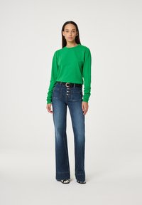 Groen langemouwen top met schouderknopen, gecombineerd met hoge taille blauwe flared jeans met voorknopen en zakken, gedragen met gepatteerde schoenen.