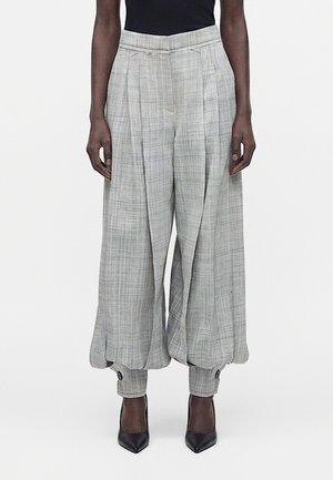 Personne grande portant un pantalon gris à carreaux taille haute, jambes larges, avec des poignets froncés et des talons noirs pointus.