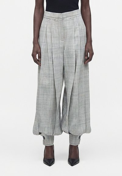 Personne grande portant un pantalon gris à carreaux taille haute, jambes larges, avec des poignets froncés et des talons noirs pointus.