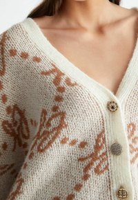 Cardigan de punto texturizado en color crema con patrones abstractos marrones. Presenta un escote en V profundo y tres botones decorativos.