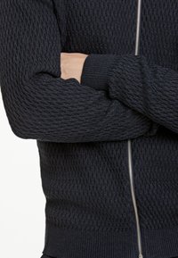 Suéter de punto texturizado azul marino con cierre frontal, puños y dobladillo acanalados. El material presenta un patrón de diamante en toda la tela.