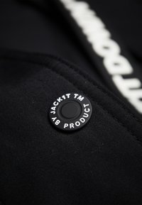 Černá látka s matným povrchem má kruhový černý patentní knoflík s bílým vystouplým textem "JACK1T TM BY PRODUCT."