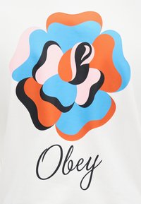 Λευκό ύφασμα με μεγάλο, πολύχρωμο σχέδιο λουλουδιού σε μπλε, ροζ, πορτοκαλί και μαύρο, με τη λέξη "Obey" σε κομψή μαύρη γραφή.