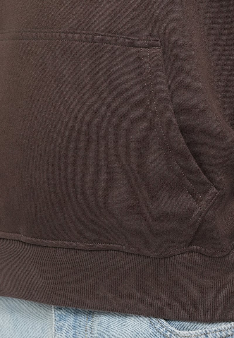 Brauner Baumwoll-Sweatshirt mit einer Vordertasche, geripptem Saum und glatter Textur, kombiniert mit heller blauer Jeans.