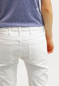 Vaqueros de mezclilla blancos con bolsillos traseros, de corte ajustado y superficie texturizada, combinados con una parte superior azul jaspeada que muestra la cinturilla.