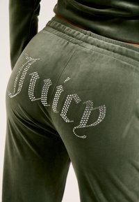 Zelené sametové joggers s rebrovaným pásom a aplikáciou rhinestonov, ktorá na zadnej strane vypisuje "Juicy", s hladkou a jemnou textúrou.