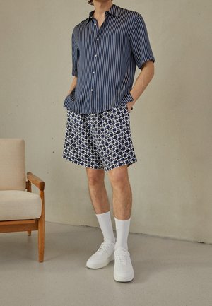 Mand iført en marineblå kortærmet skjorte med lodrette striber, mønstrede navyblå og hvide shorts, hvide sokker og hvide sneakers, stående indendørs.