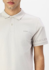 Hellbeige Polo-Shirt mit Kragen, ausgestattet mit zwei Knöpfen und dezentem "GUESS" Logo-Stick auf der Brust und dem Ärmel. Aus Baumwollmaterial.