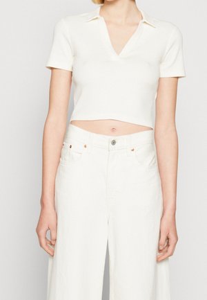 Persoon die een wit kort mouwcropped top met kraag en hoog getailleerde witte broek draagt tegen een effen achtergrond.