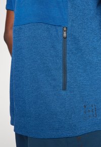 T-shirt atlético azul com um acabamento texturizado, apresentando um bolso com fecho e um detalhe de texto impresso na parte inferior. Mangas curtas e design de cor sólida.
