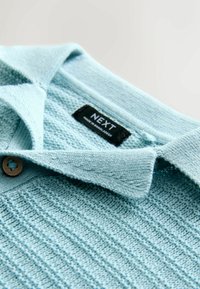 Világoskék kötött galléros póló texturált mintával, gombbal és egy fekete címkével, amin az áll: "NEXT". Galléros kialakítás.