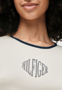 Κοντομάνικη κρέμα μπλούζα με ναυτίες ριγέ λαιμόκοψη και μπλε κεντητό λογότυπο "HILFIGER" κεντραρισμένο στο στήθος. Λείο υφασμάτινο υλικό.
