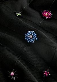 Černá texturovaná látka s barevnými korálkovými ozdobami v modré, růžové a zelené barvě, uspořádanými do květinových vzorů.