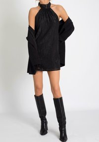 Vestido negro tipo halter con una textura brillante, que presenta un escote fruncido. Combinado con una chaqueta negra de gran tamaño y botas de cuero hasta la rodilla.