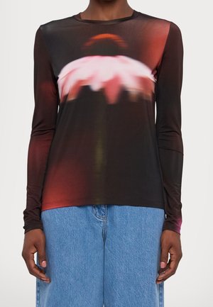 Person trägt ein langärmeliges schwarzes Oberteil mit einem verschwommenen Blumenmuster in Pink und Rot, kombiniert mit hellblauen Jeans.
