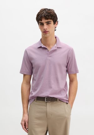 Junger Mann trägt ein hellviolettes kurzärmliges Poloshirt, das in beige Hose mit schwarzem Gürtel gesteckt ist, und steht vor einem schlichten weißen Hintergrund.