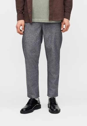 Nedre del av en person som har på seg grå bukser, svarte blanke snøresko, lyse sokker, en grønn skjorte og en brun jakke.