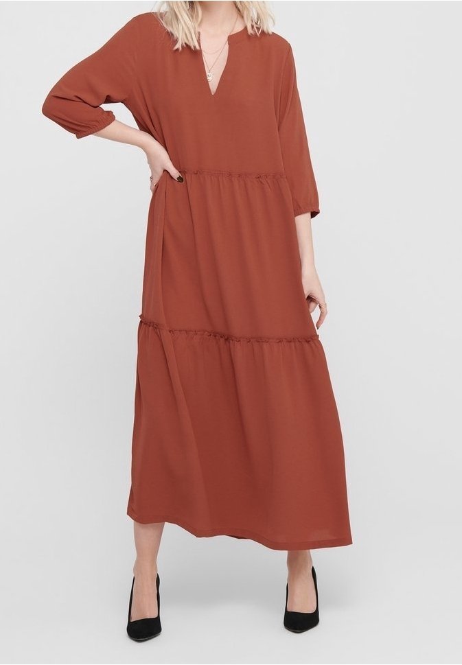 Femme portant une longue robe couleur rouille aux manches trois-quarts et des escarpins noirs, debout avec une main sur la hanche devant un fond uni.