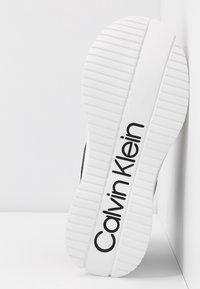 Semelle en caoutchouc blanc avec des rainures texturées, arborant le logo noir "Calvin Klein". Surface supérieure lisse en noir, présentée en angle.