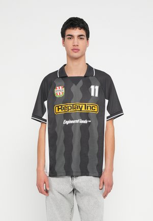 Giovane uomo con maglia sportiva a strisce ondulate nere e grigie con logo "Replay Inc", numero 11, e jeans grigi chiari, in piedi su sfondo bianco.