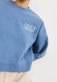 Blå bomullströja med rund halsringning, prydd med vit tryckt text "OBEY" och stadnamn. Kortad design och textil med struktur.