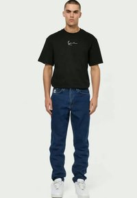 Μαύρη βαμβακερή μπλούζα με λευκό λογότυπο, σε συνδυασμό με μπλε τζιν παντελόνι. Το μοντέλο φοράει λευκά αθλητικά. Μινιμαλιστικός σχεδιασμός και άνετη εφαρμογή.