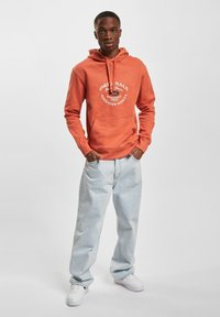 Orangene Kapuzenjacke mit vorderer Tasche und weißem Logoschriftzug, kombiniert mit hellblauen weiten Jeans und weißen Turnschuhen vor einem neutralen Hintergrund.